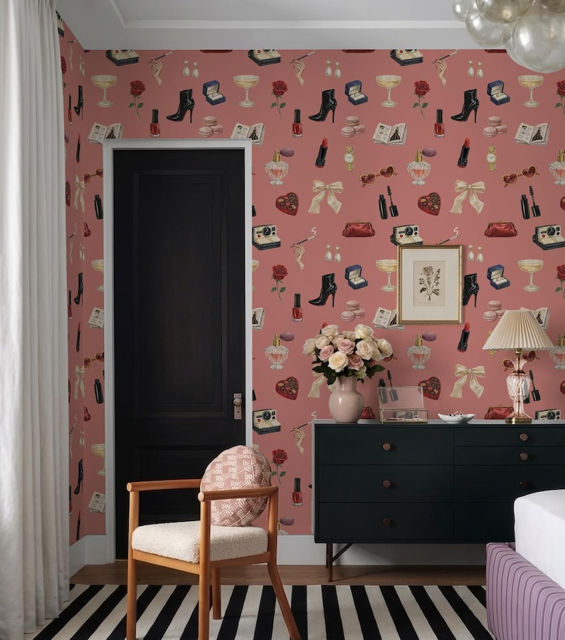 a bedroom with a pink wall adorned with a pattern of various fashion accessories, including boots, handbags, and jewelry. A black dresser with a vase of flowers and a lamp are also present in the room. A wooden chair is situated in front of the dresser, and a black door is visible on the left side of the image.