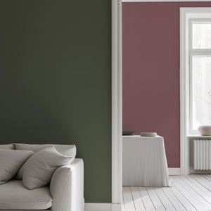 Modern interior features peel-and-stick solid color wallpapers: a deep olive green accent wall in the foreground and a muted mauve wall in the next room. A neutral sofa, whitewashed floors, and natural light complete the minimalist, elegant look.