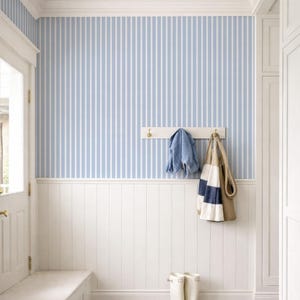May include: A hallway with blue and white striped wallpaper, white wainscoting, and a wooden floor. A white bench, door, and cabinet are visible. A denim jacket and a striped tote bag hang on a white rack. White rain boots and beige shoes are on the floor.