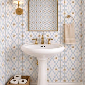 Könnte beinhalten: Badezimmerinterieur mit weißem Standwaschbecken, goldenen Armaturen und einem goldgerahmten Spiegel. Die Wände sind mit blau-gold-weiß gemusterter Tapete dekoriert. Ein Weidenkorb enthält Toilettenpapierrollen. Goldene Wandleuchte und Handtuchring.