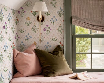 May include: A window seat with a pink tufted cushion, a solid pink velvet pillow, and a solid olive green velvet pillow. A wall-mounted lamp with a white pleated shade is attached to wallpaper featuring a floral trellis pattern. A book lies open.