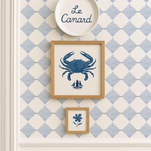 Puede incluir: Una composición mural de temática costera con un plato blanco con el texto "Le Canard", una impresión enmarcada de un cangrejo azul y una impresión enmarcada de una flor azul. Fondo de papel pintado crema con patrones de diamantes azules.