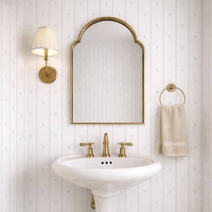 Puede incluir: Interior de baño con un lavabo blanco con pedestal y grifos dorados, un espejo con marco dorado y un toallero a juego. Una lámpara de pared con pantalla plisada está a la izquierda. El papel pintado tiene un patrón de rayas verticales con pequeños detalles florales azules.