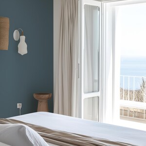 Coastal bedroom features a peel-and-stick solid color wallpaper in a muted blue, creating a calming focal point. Neutral tones in the bedding, flowing white curtains, and a sunlit open balcony with ocean views complete the tranquil, minimalist design