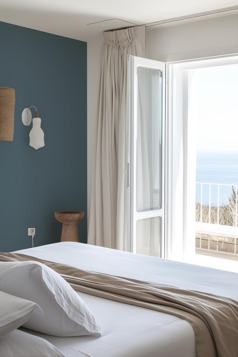 Coastal bedroom features a peel-and-stick solid color wallpaper in a muted blue, creating a calming focal point. Neutral tones in the bedding, flowing white curtains, and a sunlit open balcony with ocean views complete the tranquil, minimalist design