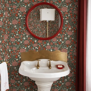 Könnte beinhalten: Badezimmer mit floraler Tapete in Grün-, Rot- und Rosatönen. Ein weißes Standwaschbecken mit Messingarmaturen steht im Mittelpunkt. Ein rot gerahmter Spiegel und eine rote Wandleuchte sind an der Wand befestigt. Ein roter Vorhang befindet sich rechts.