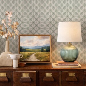 a wooden dresser with a painting, a lamp, and a vase on top. The dresser has multiple drawers and a shelf. The wall behind the dresser has a patterned wallpaper.