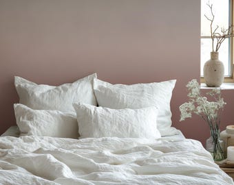 Può includere: Un set di biancheria da letto in lino bianco composto da due grandi cuscini rettangolari, due cuscini rettangolari più piccoli e un copripiumino testurizzato. Il tessuto presenta una trama visibile e un aspetto morbido e stropicciato.