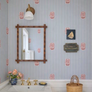 Può includere: Un bagno con carta da parati a righe blu e bianche con un motivo rosa ripetuto. Uno specchio con cornice in legno è appeso sopra un lavandino bianco con rubinetti dorati. Un piccolo cestino di vimini è appoggiato su una superficie bianca.