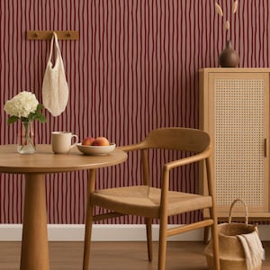Op de afbeelding: Een eethoek met een ronde houten tafel, een stoel en een kast. De achtergrond is voorzien van een bordeauxrode en lichtroze gestreepte behang. Een witte tas hangt aan een houten kapstok. Een vaas met bloemen en een fruitschaal staan op de tafel.