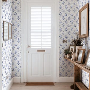 May include: A bright hallway with white walls and a white door. The walls are covered in blue floral wallpaper. A wooden table holds framed pictures and vases. The floor is light wood.