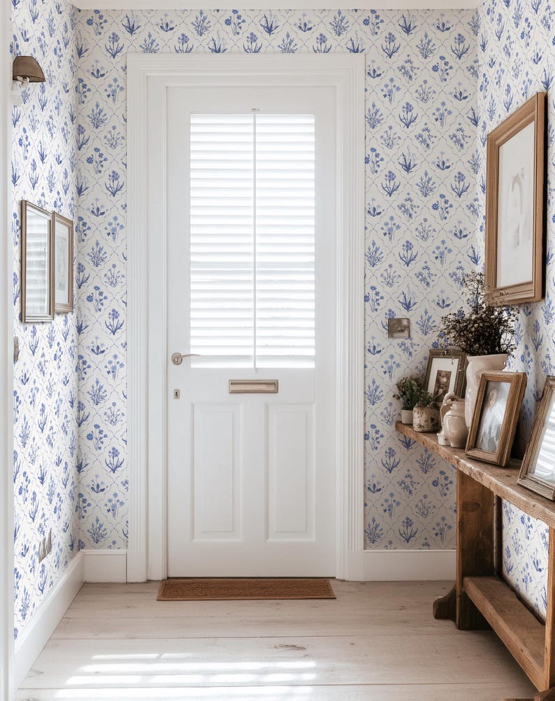 May include: A bright hallway with white walls and a white door. The walls are covered in blue floral wallpaper. A wooden table holds framed pictures and vases. The floor is light wood.