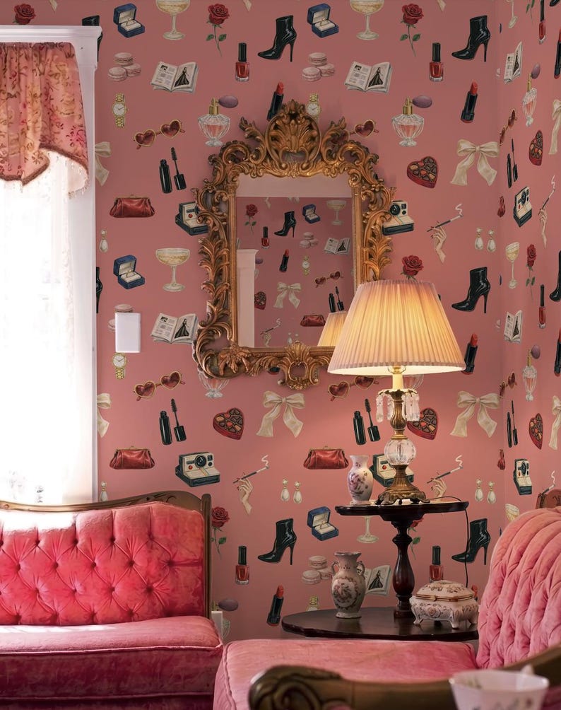 a cozy and stylishly decorated living room with a pink color scheme. The room features a pink sofa, a pink armchair, and a pink ottoman. The walls are adorned with a pink wallpaper that showcases various fashion and beauty-related icons, including high heels, makeup, and accessories. A large mirror with an ornate gold frame is mounted on the wall, and a lamp with a beige shade is placed on a small table in front of the sofa.