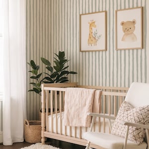 Puede incluir: Una habitación infantil con una cuna de madera clara, una mecedora blanca y una alfombra blanca y esponjosa. Las paredes están a rayas en verde claro y blanco. Dos estampados de animales en acuarela enmarcados cuelgan sobre la cuna. Una manta rosa está sobre la cuna.