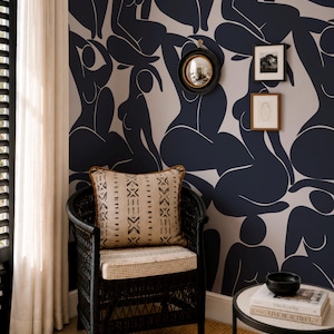 May include: A black wicker chair with a patterned cushion and pillow is positioned before a wall featuring a repeating abstract figure design in navy and white. A small side table holds books and a black bowl.