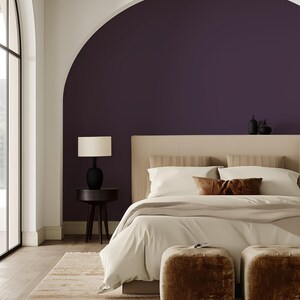 Serene bedroom with a peel-and-stick solid color wallpaper in deep plum, adding bold contrast to the arched wall niche. The design is complemented by a neutral bed, soft textures, plush ottomans, and natural light streaming through arched windows.