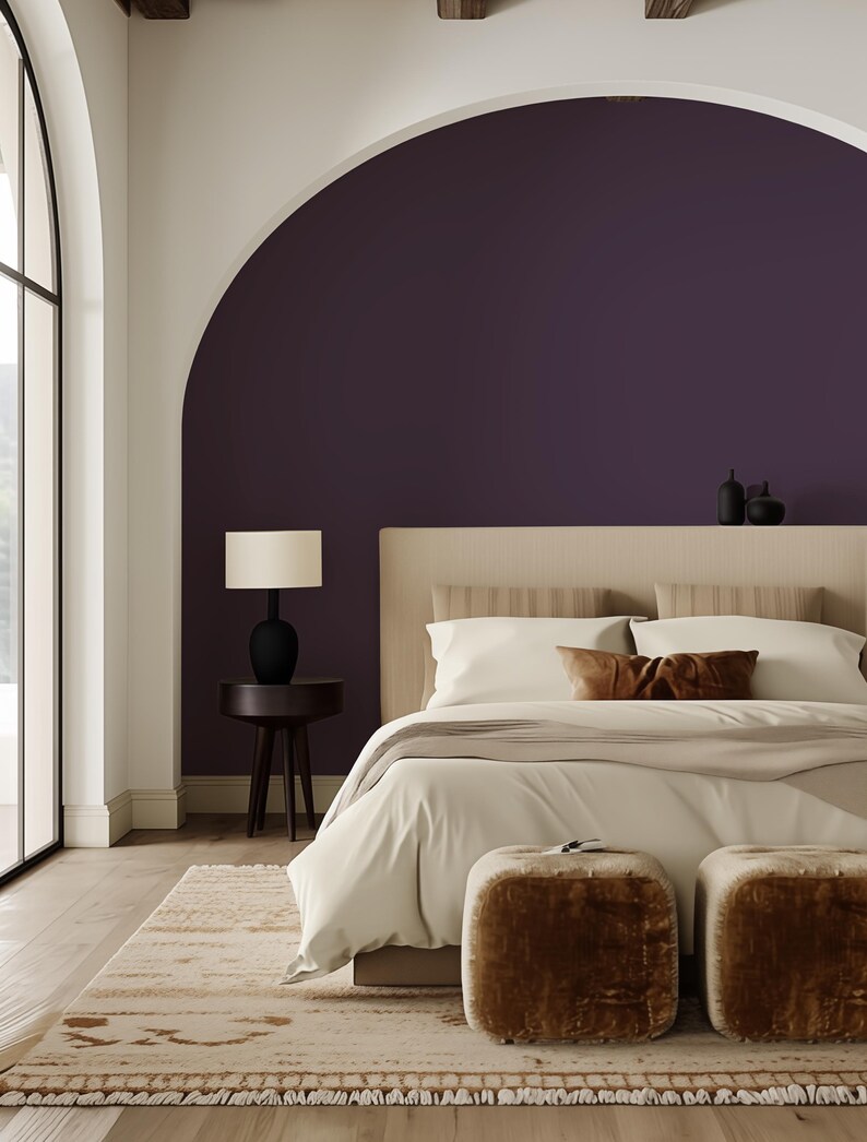 Serene bedroom with a peel-and-stick solid color wallpaper in deep plum, adding bold contrast to the arched wall niche. The design is complemented by a neutral bed, soft textures, plush ottomans, and natural light streaming through arched windows.