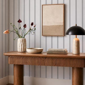 a wooden console table with a vase of flowers, a bowl, and a stack of books on it. The table is set against a striped wall, and there is a framed picture hanging on the wall.