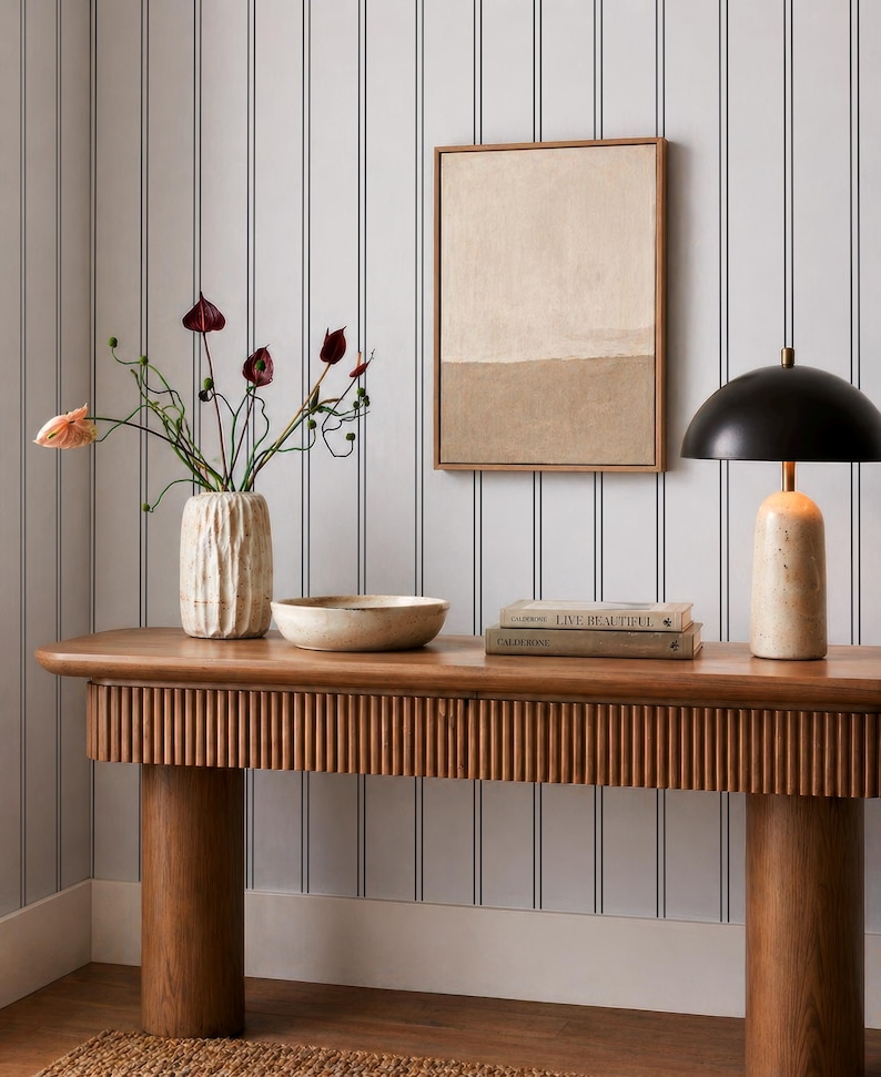 a wooden console table with a vase of flowers, a bowl, and a stack of books on it. The table is set against a striped wall, and there is a framed picture hanging on the wall.