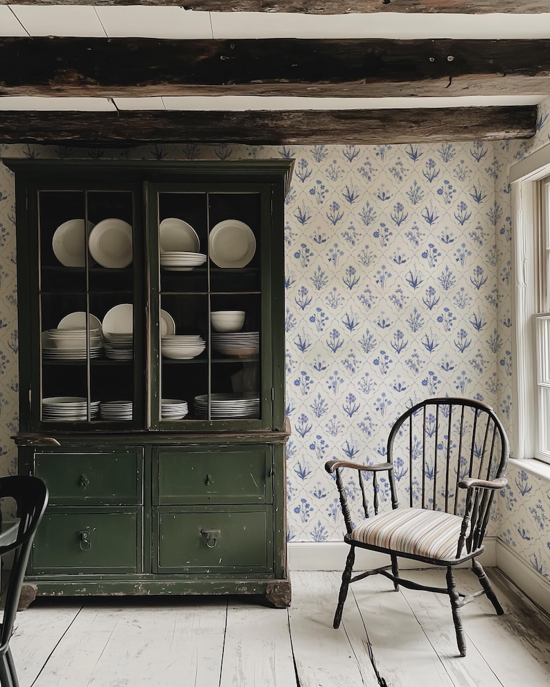 May include: A vintage-style kitchen scene featuring a dark green cabinet filled with white dishes. A black wooden chair with a striped seat sits near a floral wallpaper. The room has a rustic, farmhouse aesthetic.