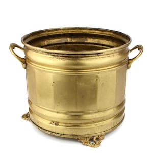 Op de afbeelding: Een gouden metalen plantenbak met twee handvatten en decoratieve poten. De plantenbak is rond en heeft een gladde, glanzende afwerking.