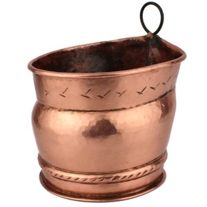 Puede incluir: Un macetero de cobre con acabado martillado y un diseño de cuerda decorativo alrededor del borde. Tiene un pequeño lazo de metal en el lateral para colgarlo.