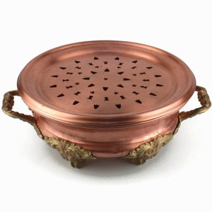 Könnte beinhalten: Eine Kupfer-Chafing Dish mit Messinggriffen und Füßen. Die Schale hat ein kreisförmiges Design mit Löchern in der Mitte.
