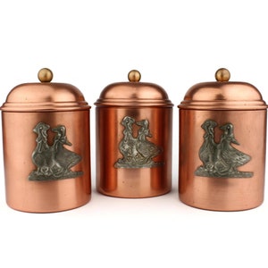 May include: Three copper canisters with lids, each featuring a silver embossed design of two ducks. The canisters are arranged in a row on a white background.
