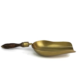 May include: A gold metal scoop with a wooden handle. The scoop is curved and has a wide, shallow bowl. The handle is dark brown and has a smooth finish.