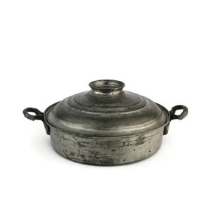 Op de afbeelding: Een zilverkleurige metalen pot met een deksel en twee handvatten. De pot heeft een versleten, verweerde uitstraling.