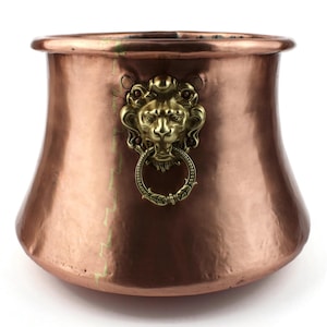 Peut inclure: Un pot en métal de couleur cuivre avec une poignée décorative en forme de tête de lion en laiton. Le pot a une forme évasée et une base arrondie. La tête de lion a un anneau dans la bouche.