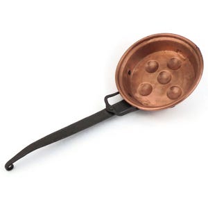 May include: Copper escargot pan with six circular indentations. The pan has a dark metal handle with a loop at the end. The pan is made of copper and has a shiny, reflective surface. The handle is attached to the pan with a small metal bracket.