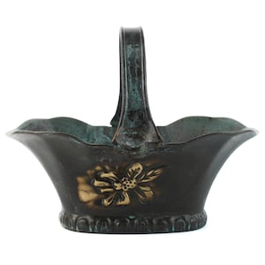 Può includere: Un cestino in metallo color bronzo scuro con un unico manico arcuato, un bordo smerlato e un rilievo floreale dorato sul davanti. La superficie presenta una patina invecchiata con ossidazione verde-azzurra visibile nelle fessure.