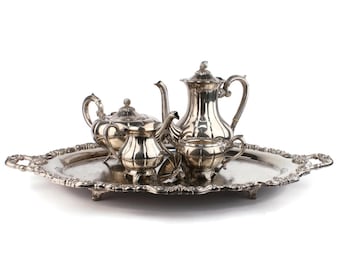May include: A silver tea set, including a teapot, creamer, sugar bowl, and cups, sits on a decorative silver tray. The set has ornate detailing and a reflective surface, perfect for serving tea or coffee. The tray has a scalloped edge.