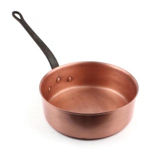 May include: A copper saucepan with a dark, wrought iron handle. The pan has a rounded base and a slightly flared rim. Rivets secure the handle to the pan. The handle has a loop for hanging. The pan is designed for cooking.