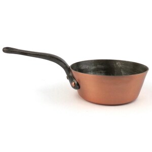 May include: A small copper saucepan with a dark, curved handle. The pan has a rounded bowl shape and a riveted handle. The interior of the pan has a dark, slightly textured finish. The handle is made of a dark metal.