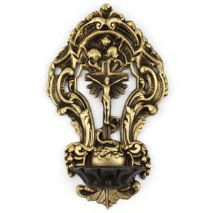May include: Ornate brass wall-mounted holy water font featuring a crucifix with a figure of Jesus. The design includes detailed scrollwork and a small basin at the bottom. The overall color is a warm gold with dark accents.