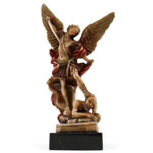 May include: A hand-painted resin figurine of Saint Michael the Archangel with large wings, wearing Roman-style armour and a red cape, holding a sword while standing over a defeated figure, all mounted on a solid black rectangular base.