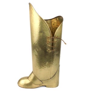 May include: A tall, golden-colored metal boot-shaped vase. The vase has a textured surface and a decorative lace-up detail on the side. The boot's opening has a unique, flared design. A decorative piece for home decor.