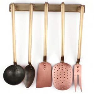 May include: A set of five kitchen utensils hanging on a brass bar. The utensils include a ladle, a spoon, a spatula, a slotted spoon, and a fork. The handles are gold-colored, and the working ends are copper-colored.