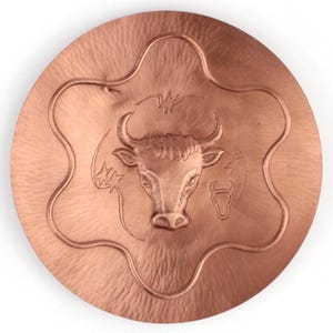 May include: A copper-coloured, round metal plate with a six-pointed star design around the edge. The centre features a detailed relief of a bull's head, with horns and facial features. The plate has a hammered texture.