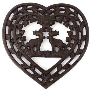 Puede incluir: Un salvamanteles de hierro fundido en forma de corazón. El diseño detallado presenta dos ciervos enfrentados bajo un árbol, rodeados de hojas. El color es marrón oscuro.