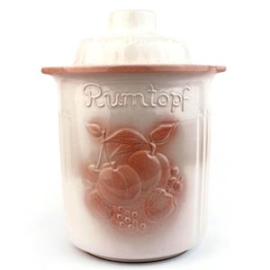 May include: A white ceramic Rumtopf jar with a lid. The jar features a raised relief design of fruit, including cherries, apples, and grapes, in a reddish-brown colour. The word "Rumtopf" is embossed on the front.