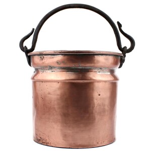 Puede incluir: Un cubo de cobre con un asa de hierro forjado oscuro. El cubo tiene forma cilíndrica con un borde ligeramente ensanchado. El cobre tiene un tono marrón rojizo cálido, con zonas más oscuras. El asa es curva y se sujeta con lazos decorativos.