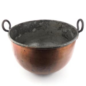 Peut inclure: Un bol en cuivre avec deux poignées en boucle. Le bol a une forme arrondie avec un intérieur sombre et un extérieur poli. Les poignées sont sombres et semblent être en fer.