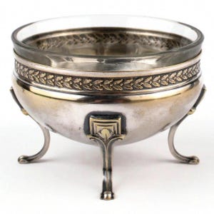 May include: A silver-colored, three-legged bowl with a clear glass insert. The bowl features a decorative band with a gold-colored leaf pattern. The legs have a curved design, and the bowl has a classic, elegant appearance.