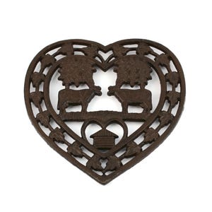 Peut inclure: Un dessous de plat en fonte de fer en forme de cœur de couleur marron avec un motif de deux vaches sous des arbres et une petite maison au centre.