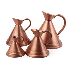 May include: Set of four copper pitchers of varying sizes. The pitchers have a conical shape with a flared lip and a handle.