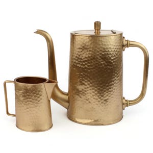 May include: A gold-coloured hammered metal coffee pot and creamer set. The coffee pot features a long, curved spout, a handle, and a lid with a decorative knob. The creamer has a handle and a spout. The set is made of metal.