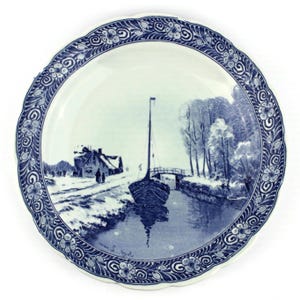 May include: A round, white ceramic plate with a blue and white landscape scene. The plate features a boat on a canal, a bridge, and a house. The rim has a scalloped edge and a floral and scroll design.